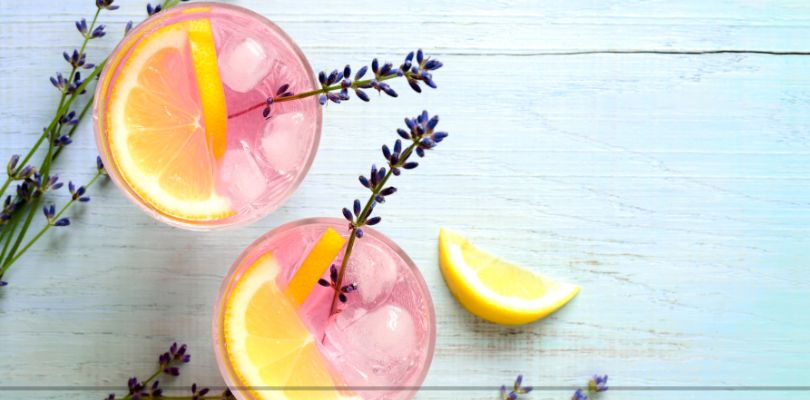 Two pink drinks with slices of lemon and sprigs of lavender in them on top of a light blue table.